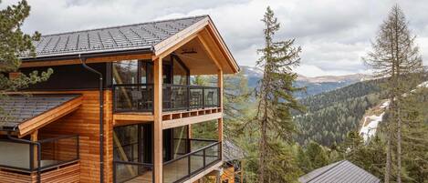 A two-story wooden cabin with large windows and a balcony, surrounded by forest and mountains.