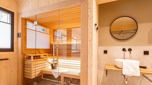 Sauna, jossa on puiset lauteet ja lasiovi, puinen penkki, jossa on pyyhe, peilillä varustettu pesuallas ja ikkuna, jossa on sälekaihtimet.