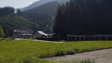 A rural setting with a building that appears to be a guesthouse or small hotel, surrounded by lush greenery and a mountain in the background.