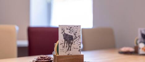 Una mesa de madera con un mantel individual, un pequeño bloque de madera con una ilustración blanco y negro de un ciervo, y un tazón decorativo.