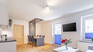Een modern appartement met een keuken, eethoek en woonruimte. Het heeft een houten eettafel, stoelen, een kleine salontafel en een aan de muur bevestigde televisie.