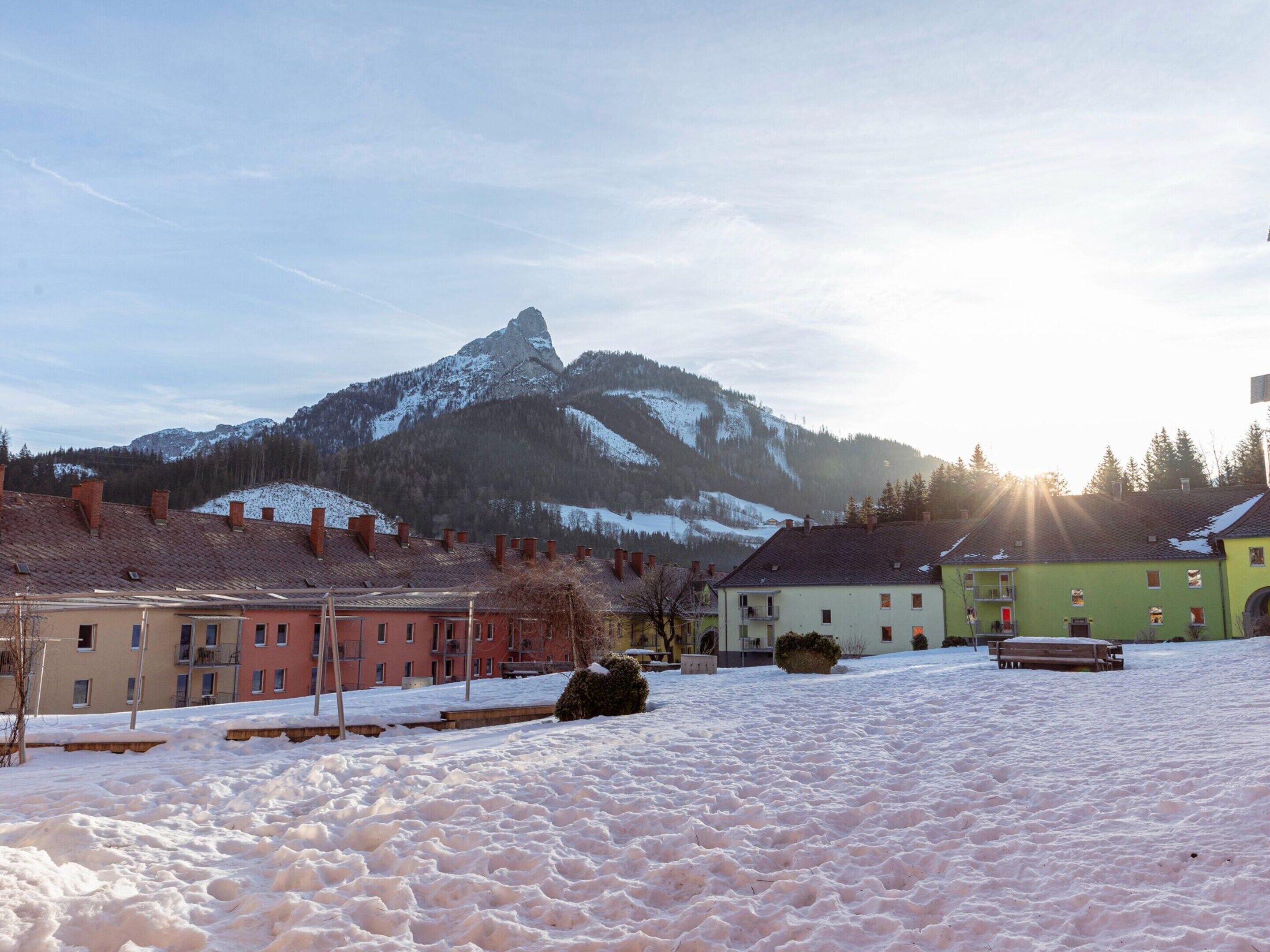 A snow-covered landscape with a mountain in the background, buildings with red and green facades, and a clear sky.