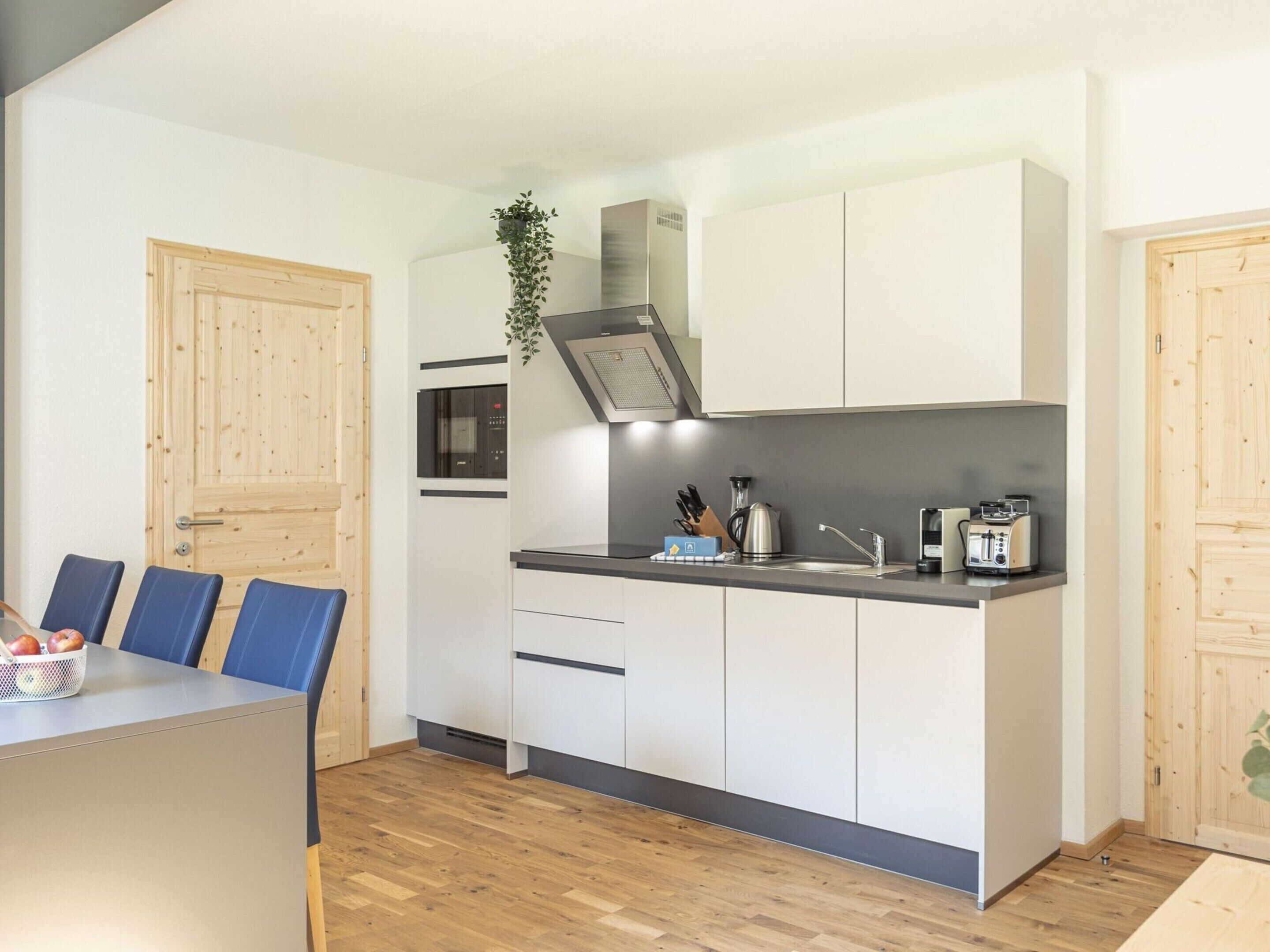 A compact kitchen with white cabinets, a built-in oven, and a sink. There is a dining table with blue chairs and a basket of apples.