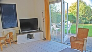 A living room with a TV on a white cabinet, a coffee table with flowers, and a wicker chair. - Bungalow Near Breskens Beach and Lighthouse (Breskens)