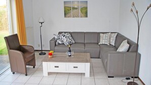 A modern living room with a grey sofa, a white coffee table, and a chair. There is a painting on the wall, a floor lamp, and a window with curtains. - Bungalow Near Breskens Beach and Lighthouse (Breskens)