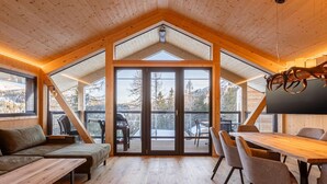A cozy cabin interior with a wooden ceiling, a dining area with chairs and a table, a sofa, and a large sliding glass door leading to a deck with a view of a snowy landscape. - Charming Chalet With hot tub and Sauna (Stadl-Predlitz)