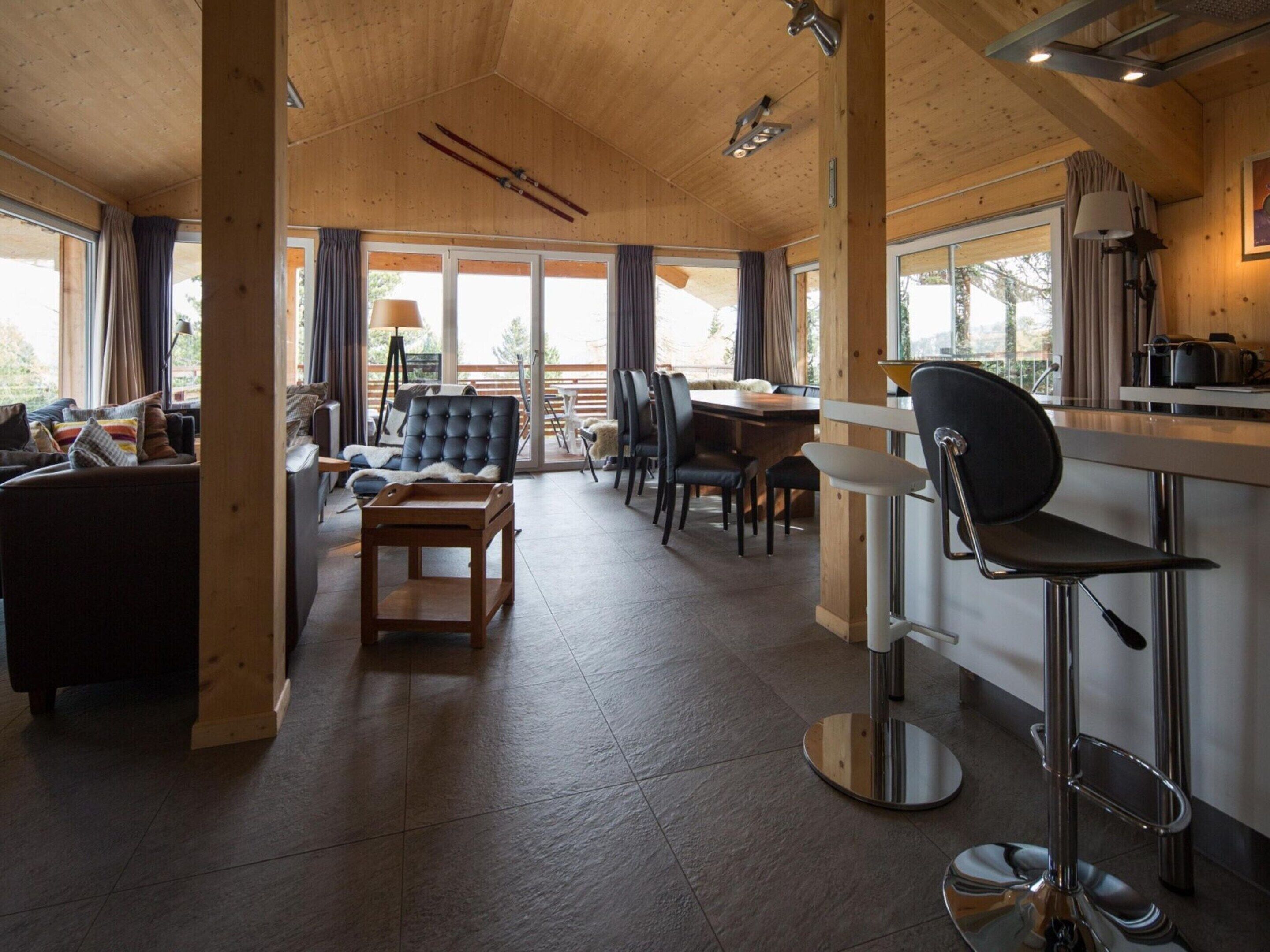 A modern interior with a wooden ceiling, a dining area with a table and chairs, a bar area with a stool, and large windows allowing natural light.