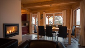 Een gezellig interieur van een blokhut met een open haard, houten plafond, een eethoek met stoelen en grote ramen met uitzicht.