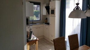 A compact kitchen with white cabinets, a microwave, and a window with blinds. There is a dining table with two chairs and a hanging light fixture. - Bungalow in Breskens Near Beach and Sea (Breskens)