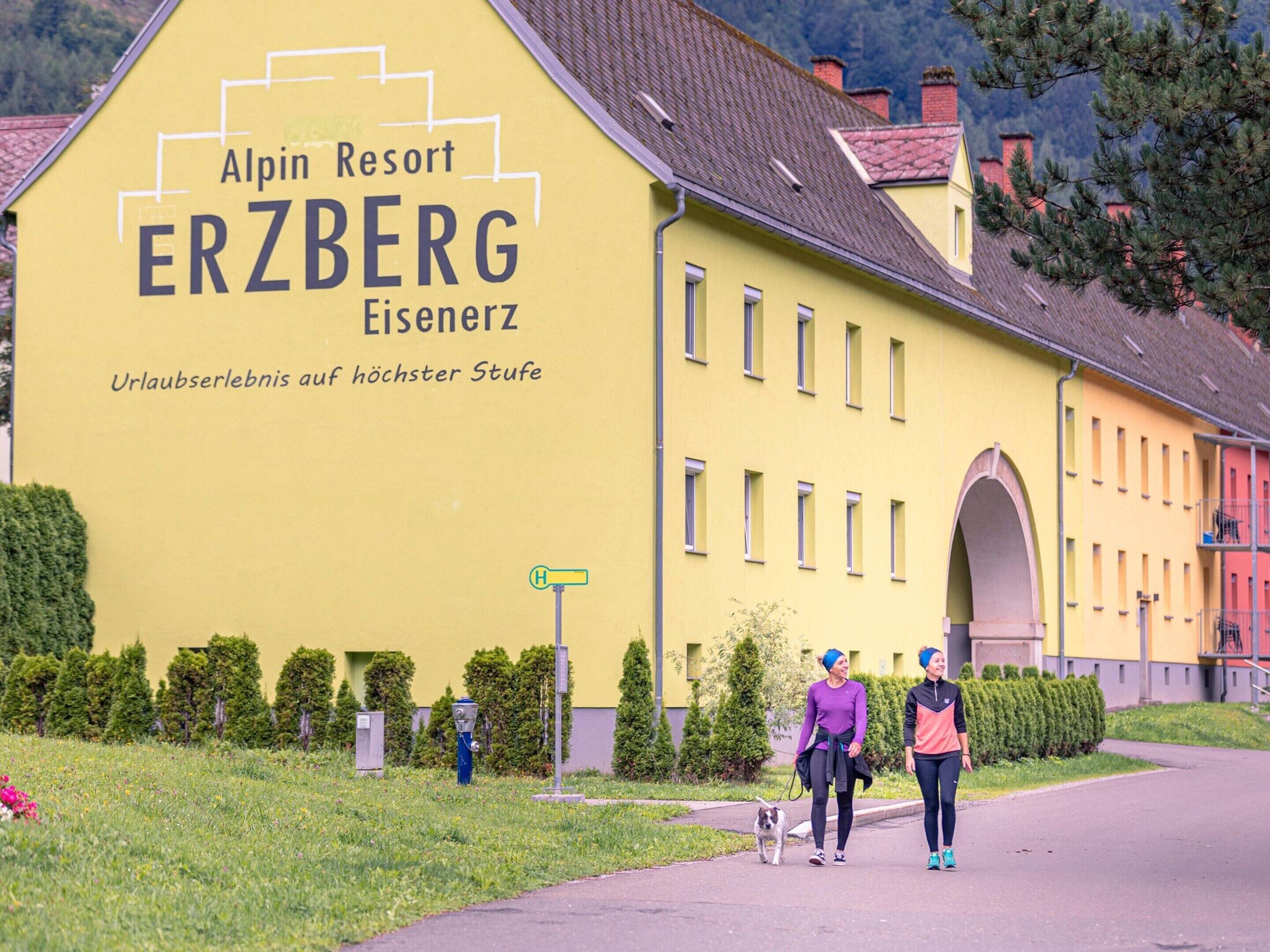 En gul byggnad med texten ”Alpin Resort ERZBERG Eisenerz” och ”En unik semesterupplevelse på högsta nivå”, vilket antyder en semesterupplevelse av högsta klass.