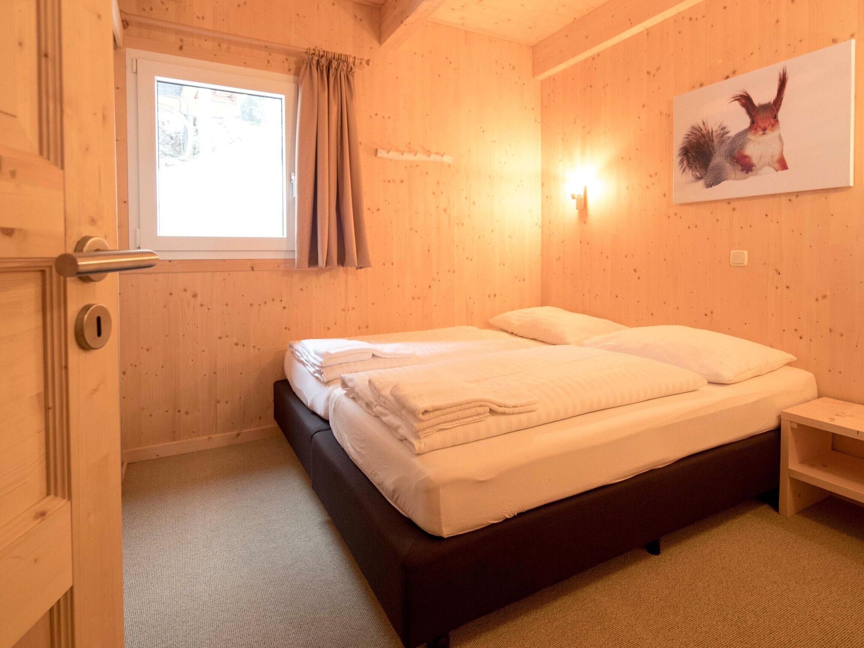 A room with a bed, a window with curtains, a door, and a wall-mounted light.