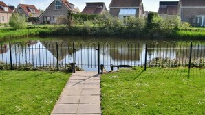 A pathway leading to a body of water with a metal fence, surrounded by greenery and residential buildings. - Bungalow in Breskens Near Beach and Pool (Breskens)