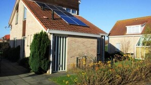 A house with a solar panel roof, a brick exterior, a small front yard with a bush and yellow flowers, and a neighboring house with a similar roof. - Bungalow in Breskens Near Beach and Pool (Breskens)