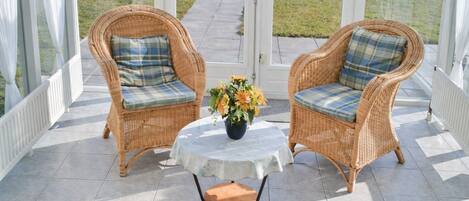 Two wicker chairs with plaid cushions, a small table with a vase of flowers, and a view of a grassy area through glass doors.