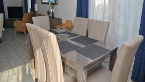 Uma sala de jantar com uma mesa retangular, seis cadeiras, uma televisão e uma janela com cortinas azuis.