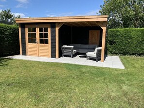 A covered outdoor seating area with a sofa and chairs, a wooden door, and a small window. - Peaceful Coastal Bungalow (Breskens)