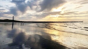 En strand med en brygga som sträcker sig ut i vattnet, en fyr i fjärran och en molnig himmel.