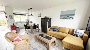 Ett vardagsrum med en soffa, ett soffbord, en fåtölj i rotting, ett matbord med stolar och ett köksområde i bakgrunden.
