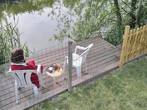 En person sitter vid ett bord på ett trädäck med utsikt över en vattenyta, och i bakgrunden syns ett staket och grönska.