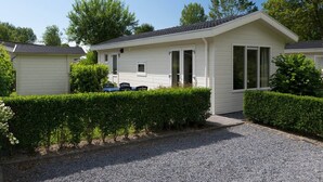 A single-story mobile home with a front porch, a small attached garage, and a gravel driveway. - House in Breskens Near the Beach (Breskens)