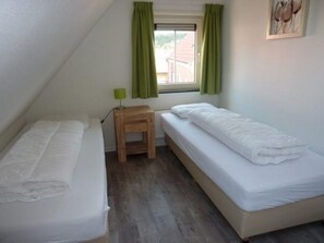 A small, single-room accommodation with two beds, a window with curtains, and a small table with a lamp. - Holiday Home Breskens Near the Coast (Breskens)