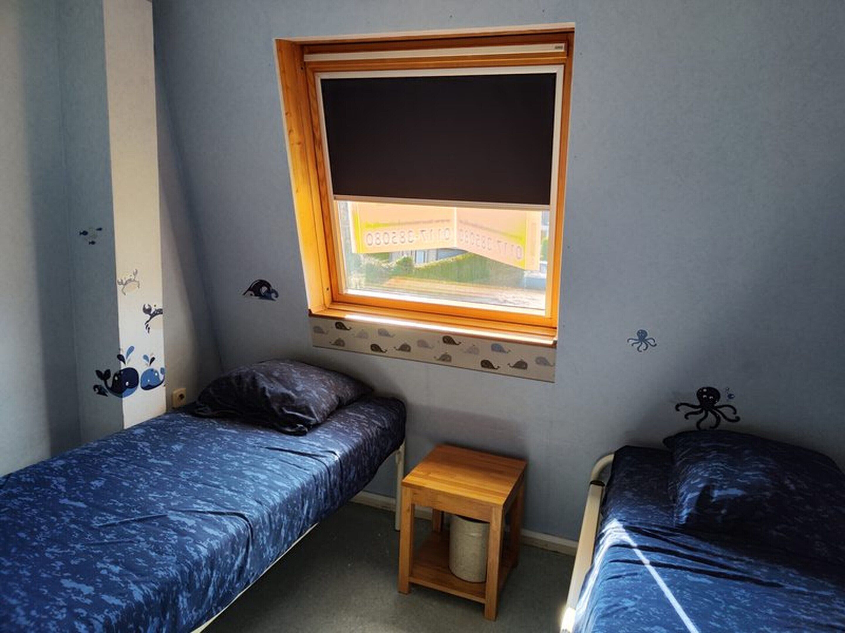 A room with two single beds, a small wooden stool, a window with a view of a building, and wall decorations.