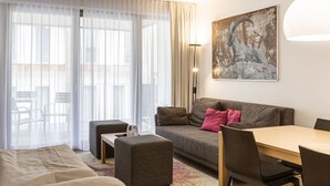 Ein modernes Hotelzimmer mit einer Couch, Hocker, Esstisch und Stühlen. Ein großes Fenster mit durchscheinenden Vorhängen, eine Stehlampe und ein Bild an der Wand sind ebenfalls vorhanden.