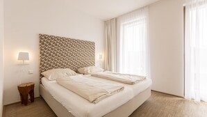 Ein modernes Hotelzimmer mit einem großen Bett, zwei Nachttischlampen, einem gemusterten Kopfteil und einem großen Fenster mit durchsichtigen Vorhängen.