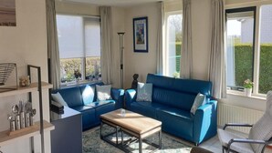 A modern living room with a blue sofa, a wooden coffee table, and a chair. There are large windows with curtains, a floor lamp, and a picture on the wall. - Detached House Breskens Near Scenic Beaches (Breskens)