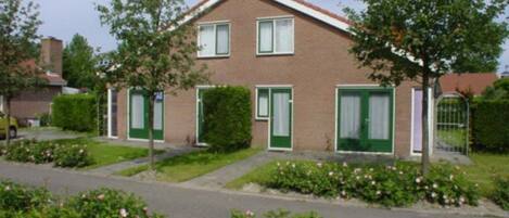 A two-story house with a brick facade, green shutters, and a small front garden with pink flowers.