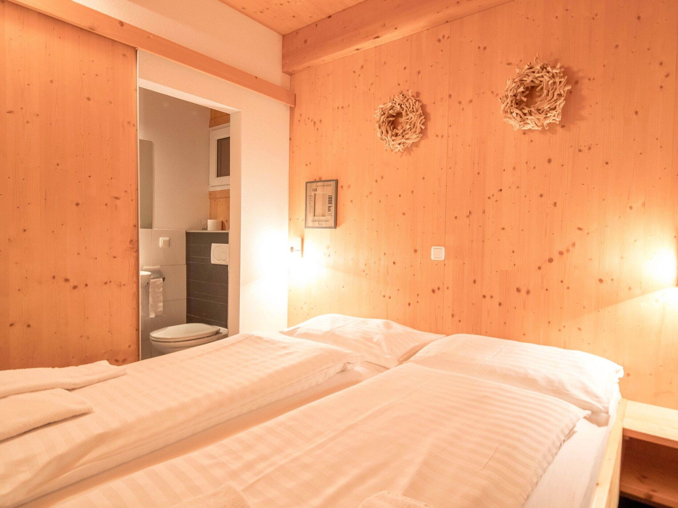 A room with wooden walls, a bed with white bedding, a small bedside table, and a wall-mounted light fixture.