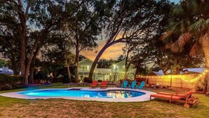 A backyard with a swimming pool, lounge chairs, and a house with string lights.