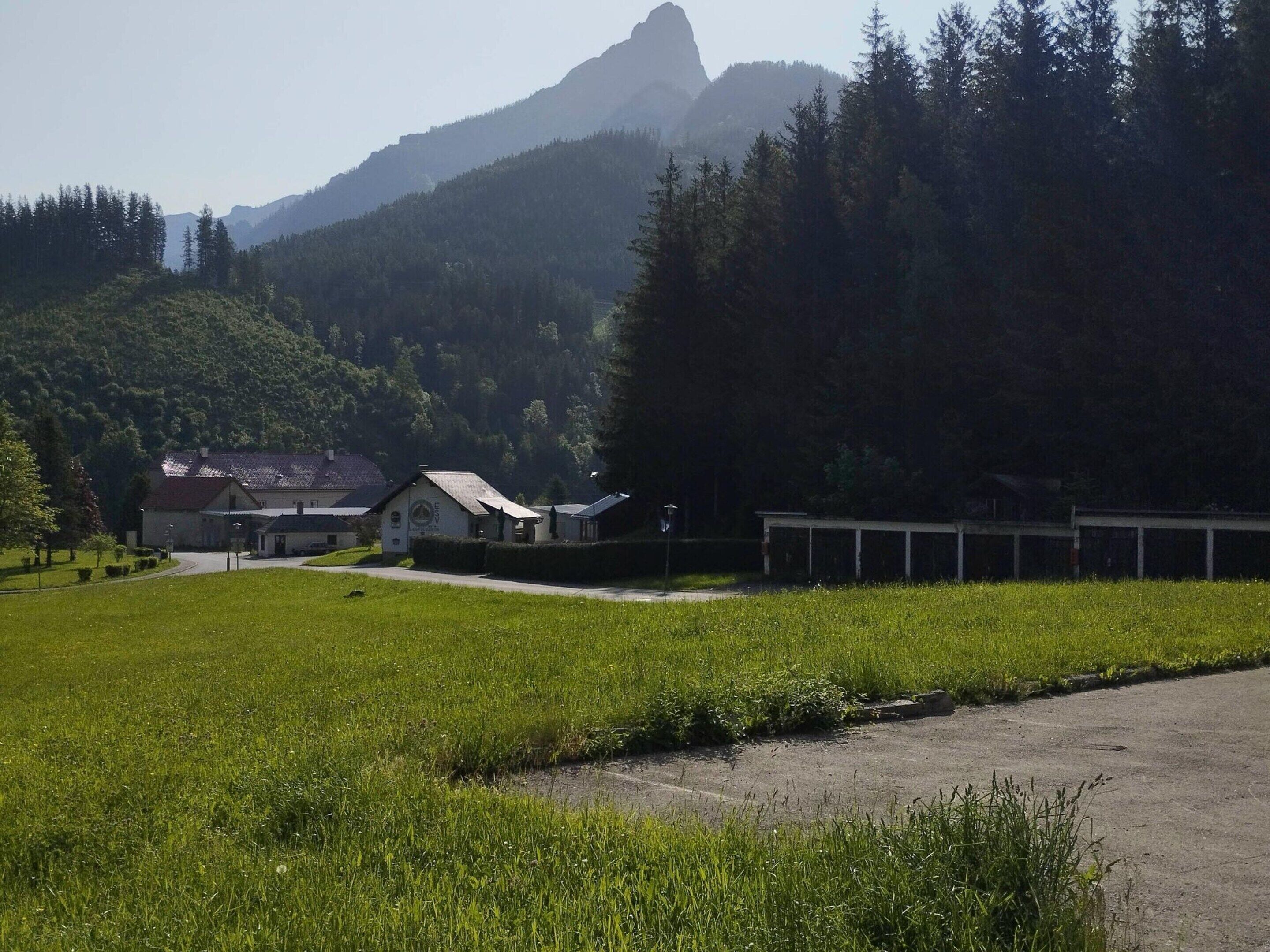 A rural setting with a building that appears to be a guesthouse or small hotel, surrounded by lush greenery and a mountain in the background.