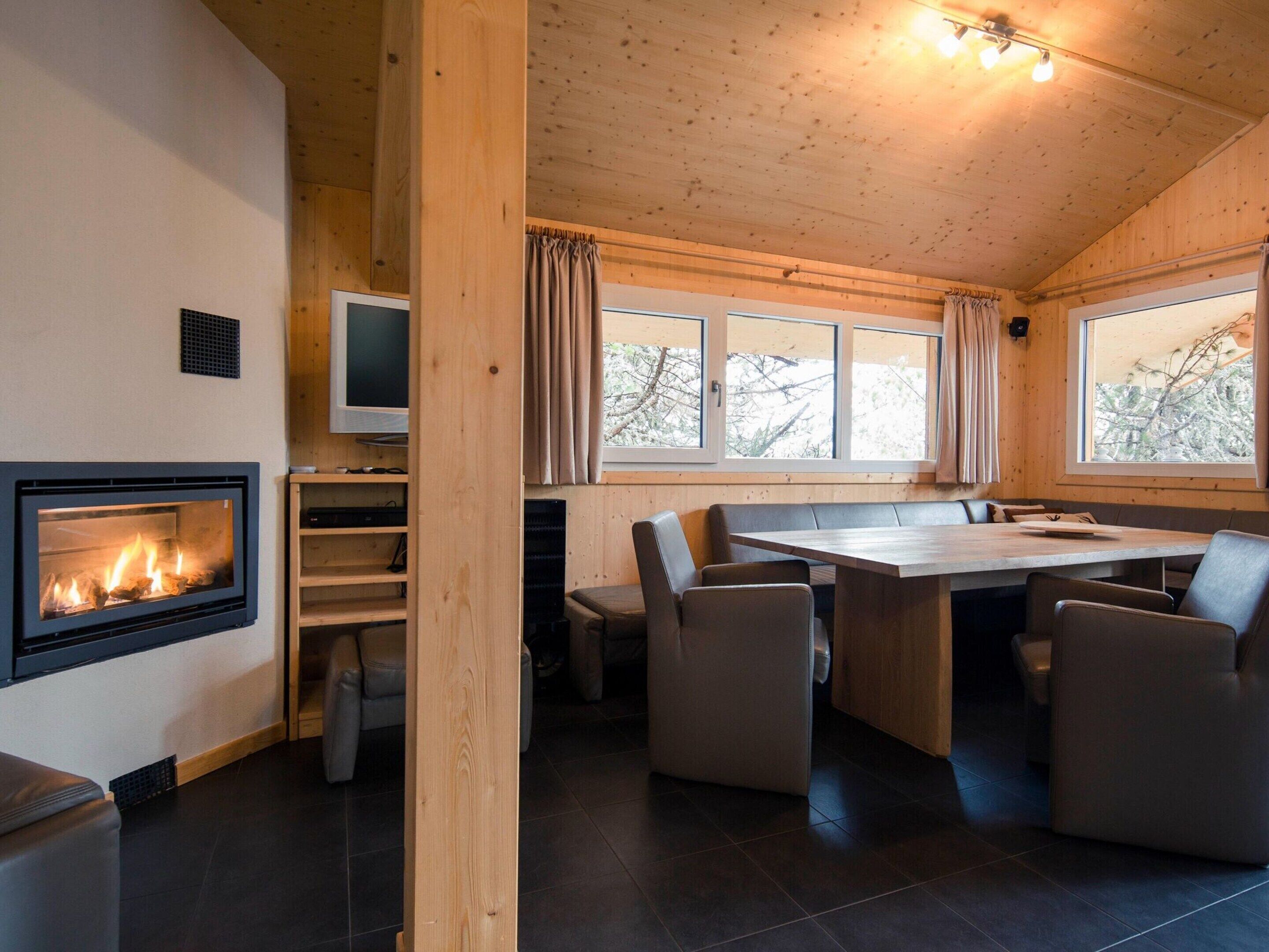 A cozy cabin interior with a fireplace, a dining table with chairs, a television, and large windows.