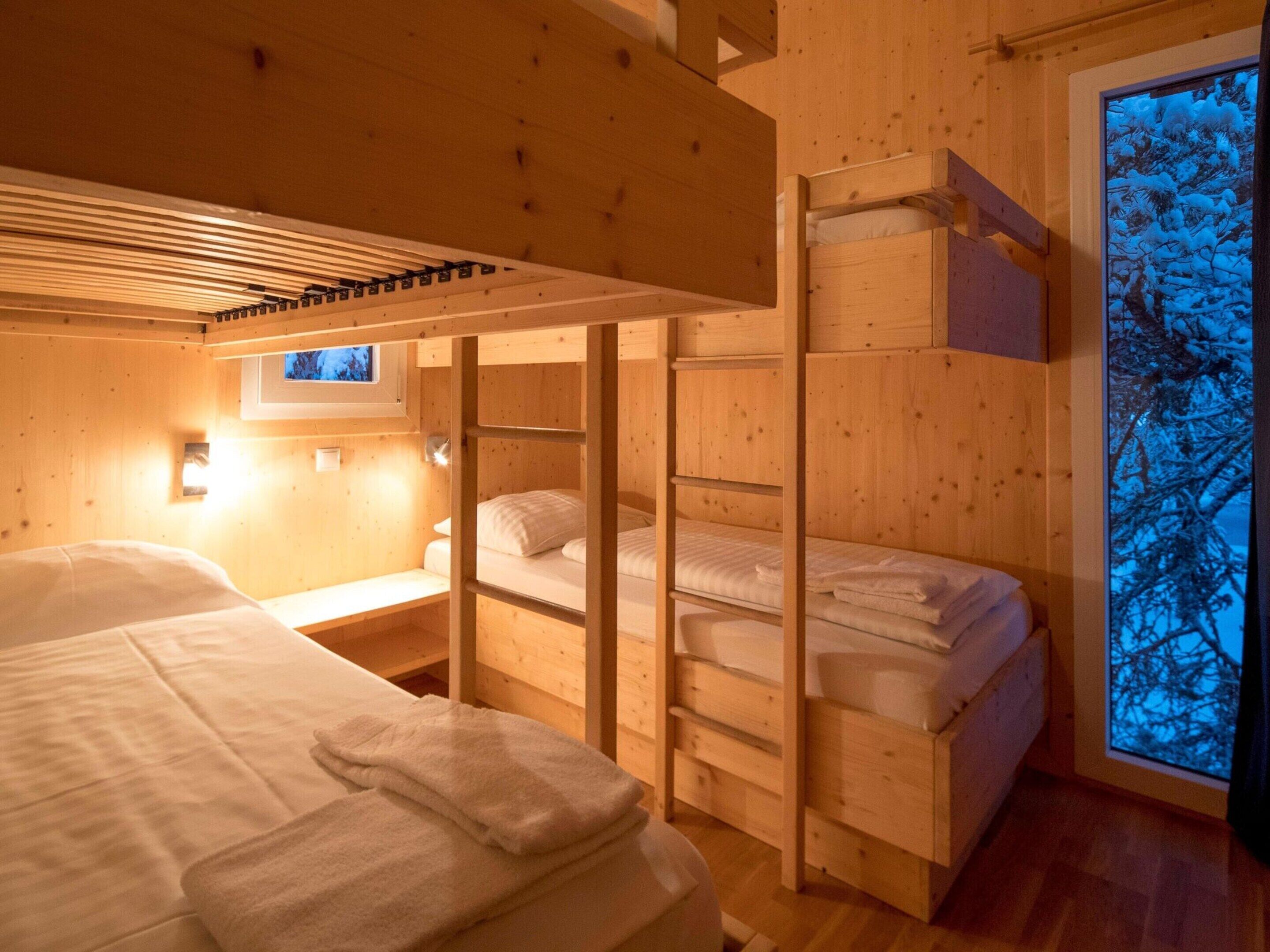 A cozy cabin with wooden walls and a large window. There are bunk beds and a small desk with a lamp.