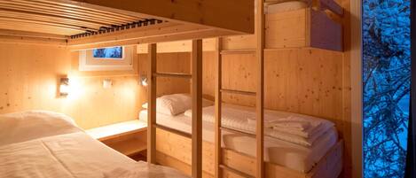 A cozy cabin with wooden walls and a large window. There are bunk beds and a small desk with a lamp.
