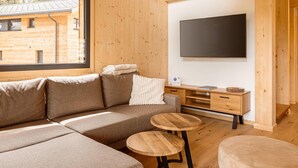 A modern log cabin interior with a sectional sofa, two wooden tables, a flat-screen TV mounted on the wall, and a window overlooking a wooden building. - Attractive Chalet Sauna & Hottub (Stadl-Predlitz)