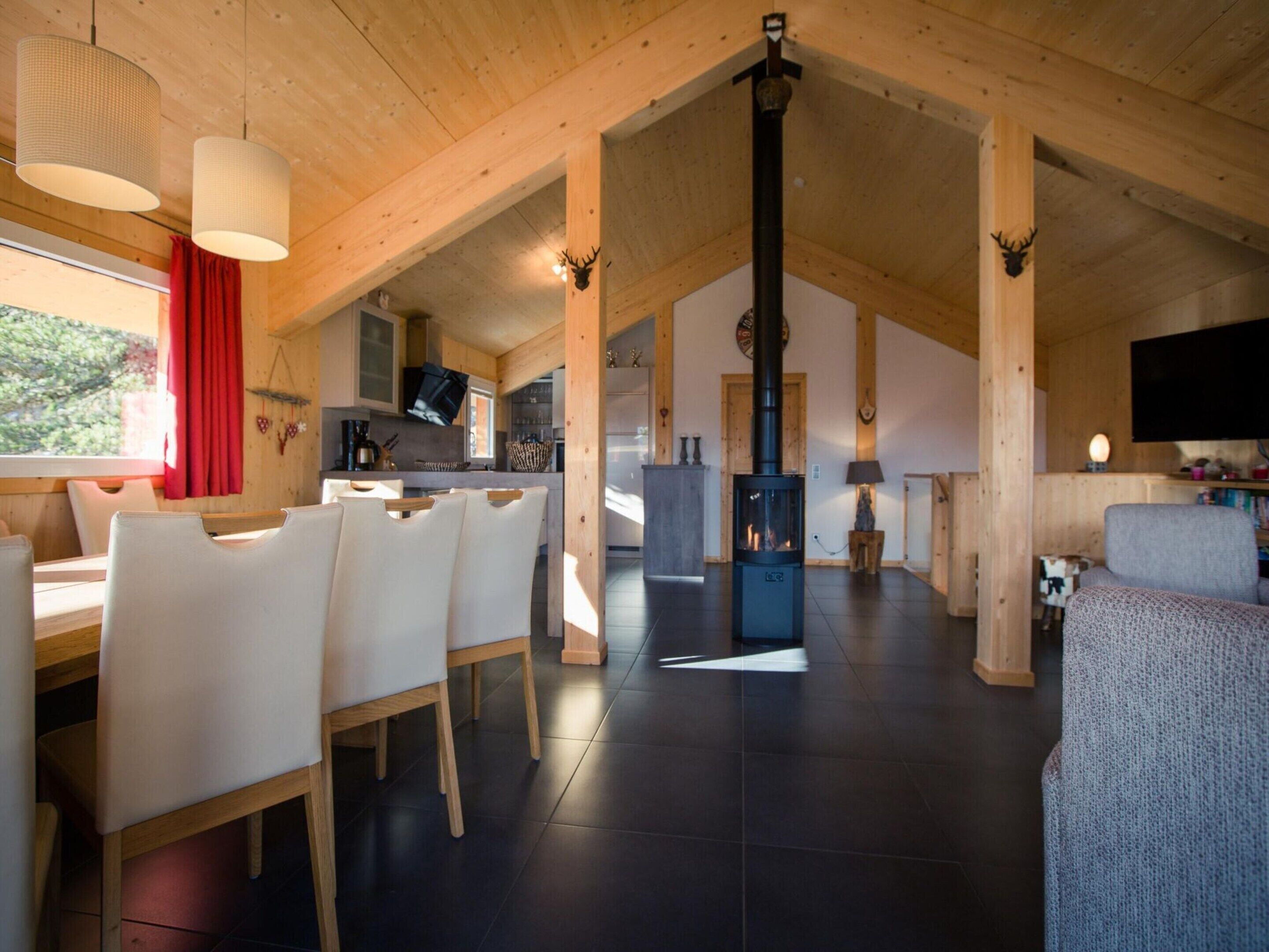 A spacious living area with a high ceiling, wooden beams, a fireplace, a dining area with white chairs, and a kitchen visible in the background.