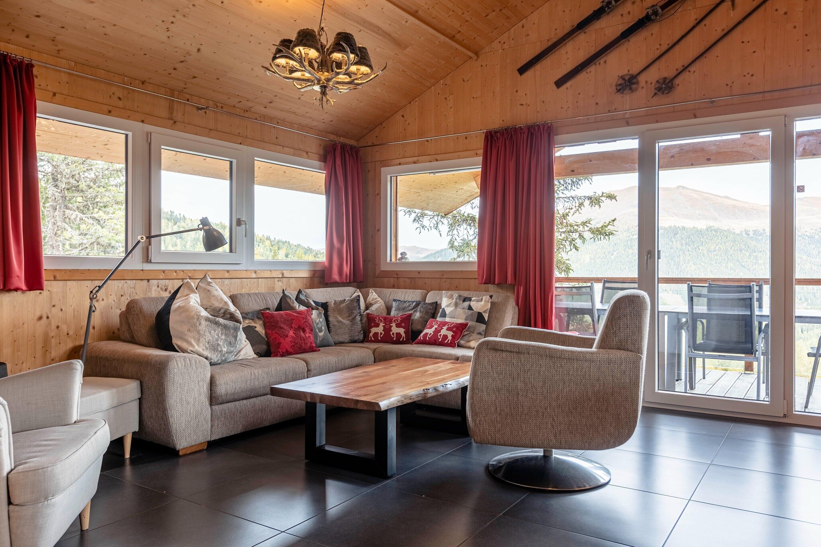 A cozy living room with a sectional sofa, a round table, and a chair. Large windows with curtains allow natural light and views of the outdoors.