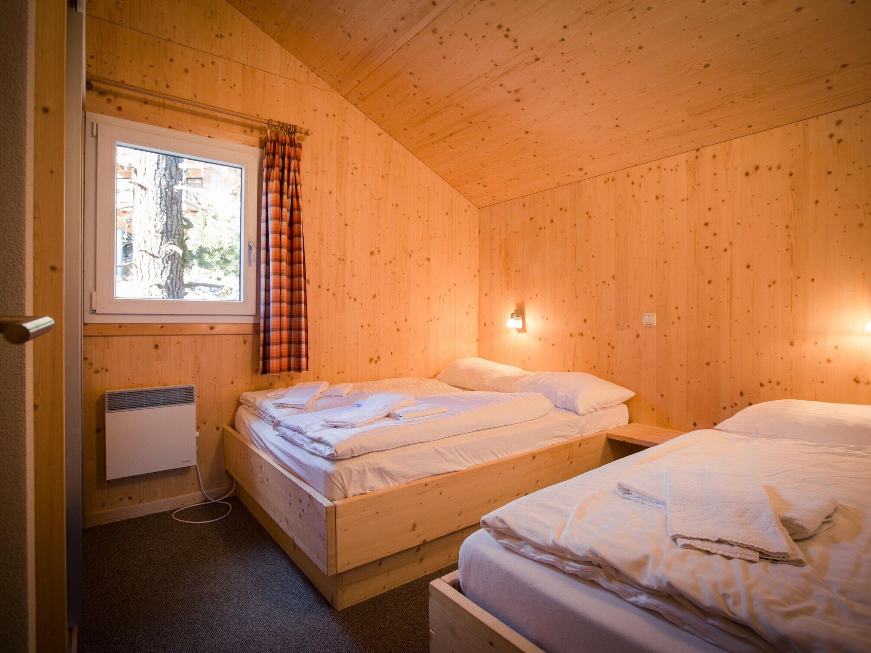 Ein Zimmer einer Holz-Blockhütte mit zwei Betten, einem Fenster mit Karomuster-Vorhang, einer Wandleuchte und einem Heizkörper.