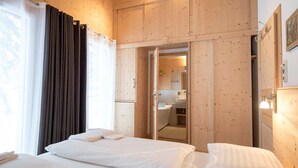 Ein Hotelzimmer mit zwei Betten, einem Holzschrank, einem durch eine offene Tür einsehbaren Badezimmer und einem mit Vorhängen versehenen Fenster.