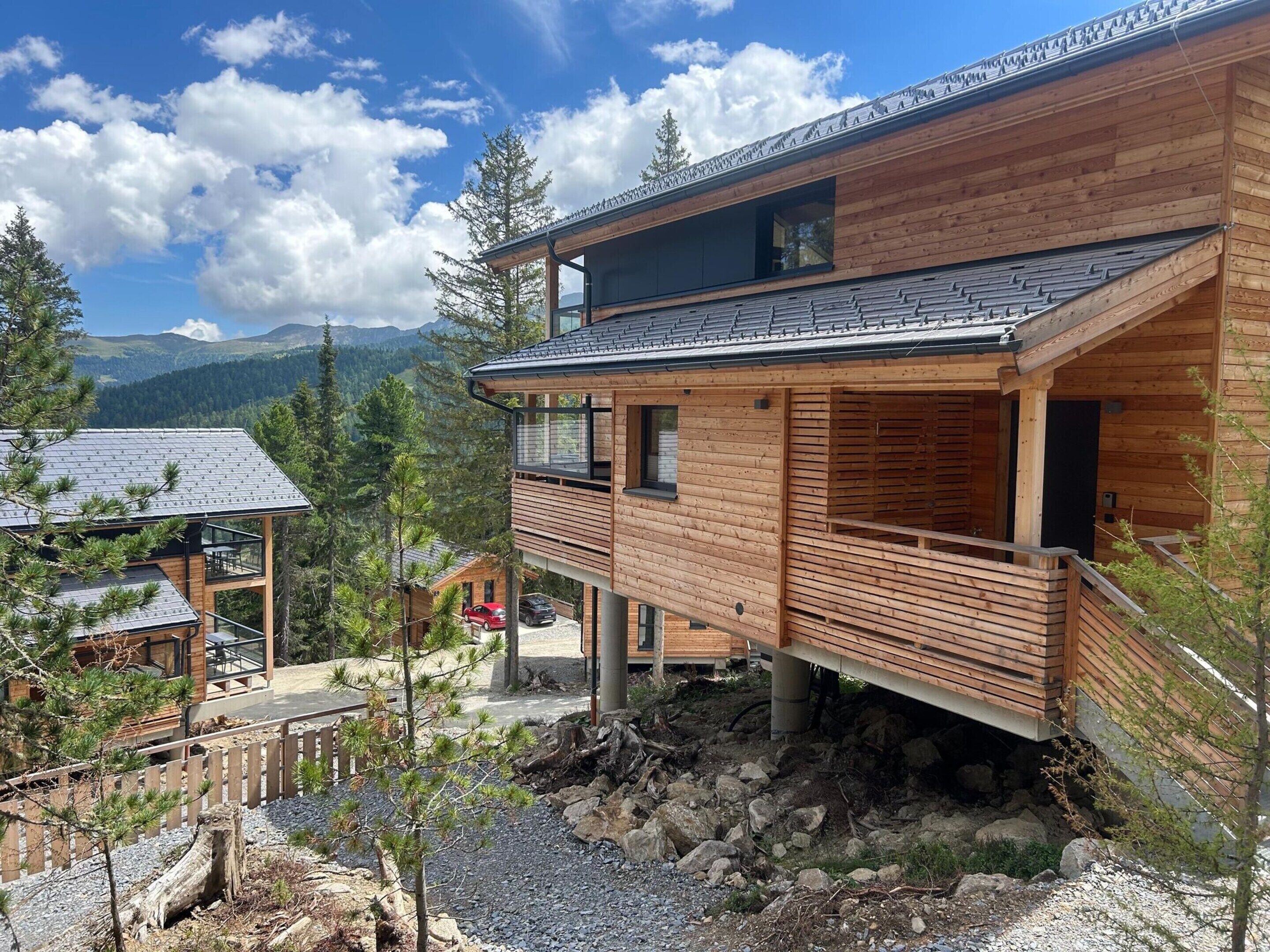A modern wooden cabin with a balcony, situated in a mountainous area with a clear sky.