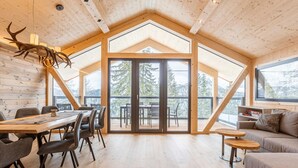 Une pièce spacieuse avec un plafond haut, des murs en bois, un coin repas avec une table et des chaises, un canapé et une grande porte-fenêtre coulissante donnant sur une terrasse extérieure d’où l’on peut admirer les arbres et les montagnes.