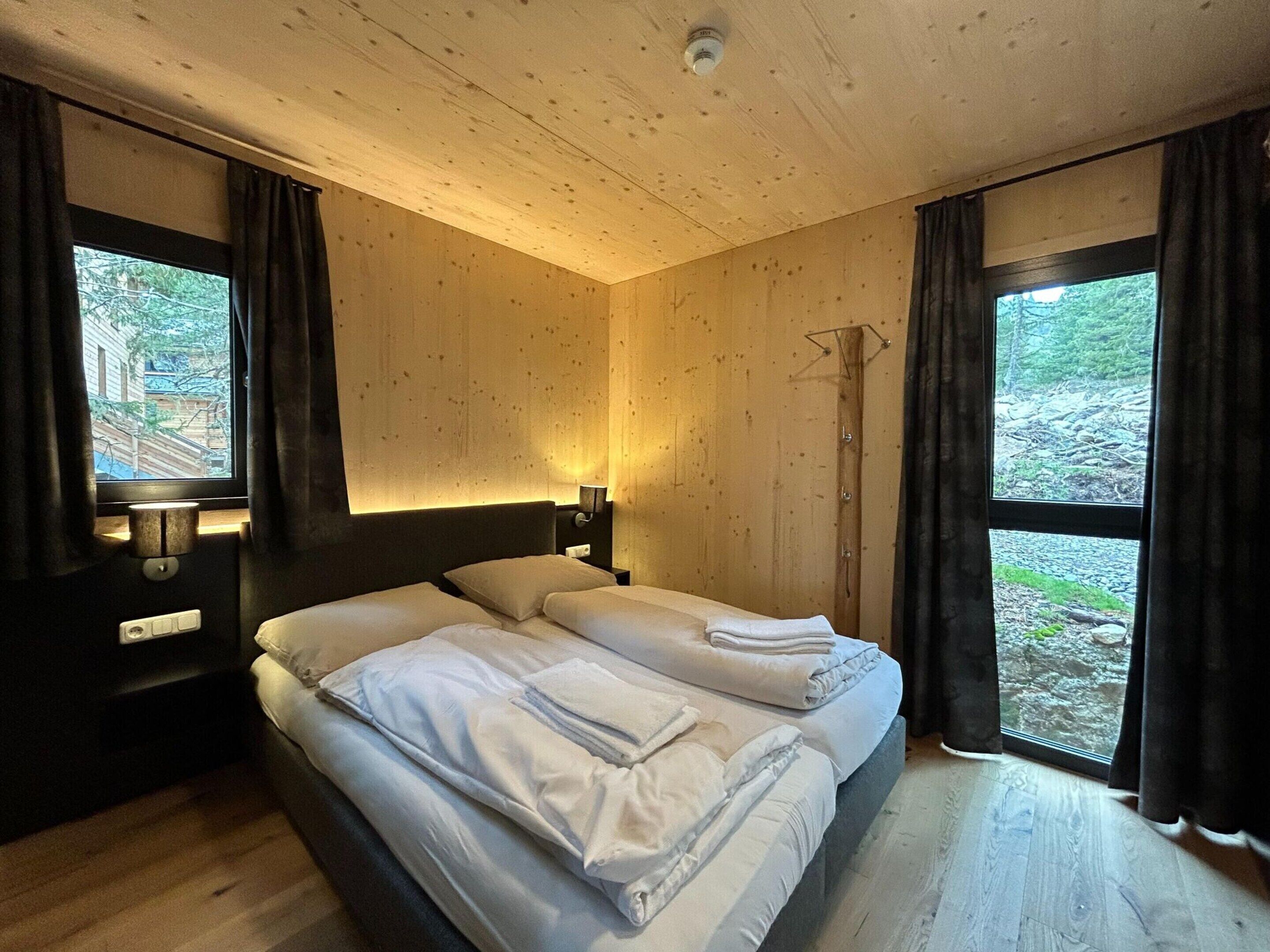 A bedroom with two beds, wooden walls, a window with curtains, and a bedside lamp.