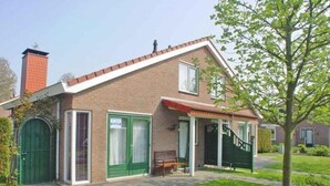 Ein eingeschossiges Haus mit Backsteinfassade, einer grünen Tür, einer kleinen Veranda mit Bank und einem Baum im Vorgarten.