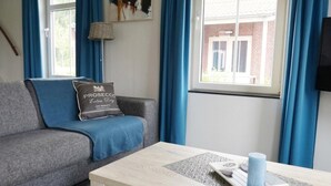 A living room with a grey sofa, a wooden coffee table, a blue cushion, a wall-mounted lamp, and a window with blue curtains. - Holiday Home in Breskens Near the Beach (Breskens)