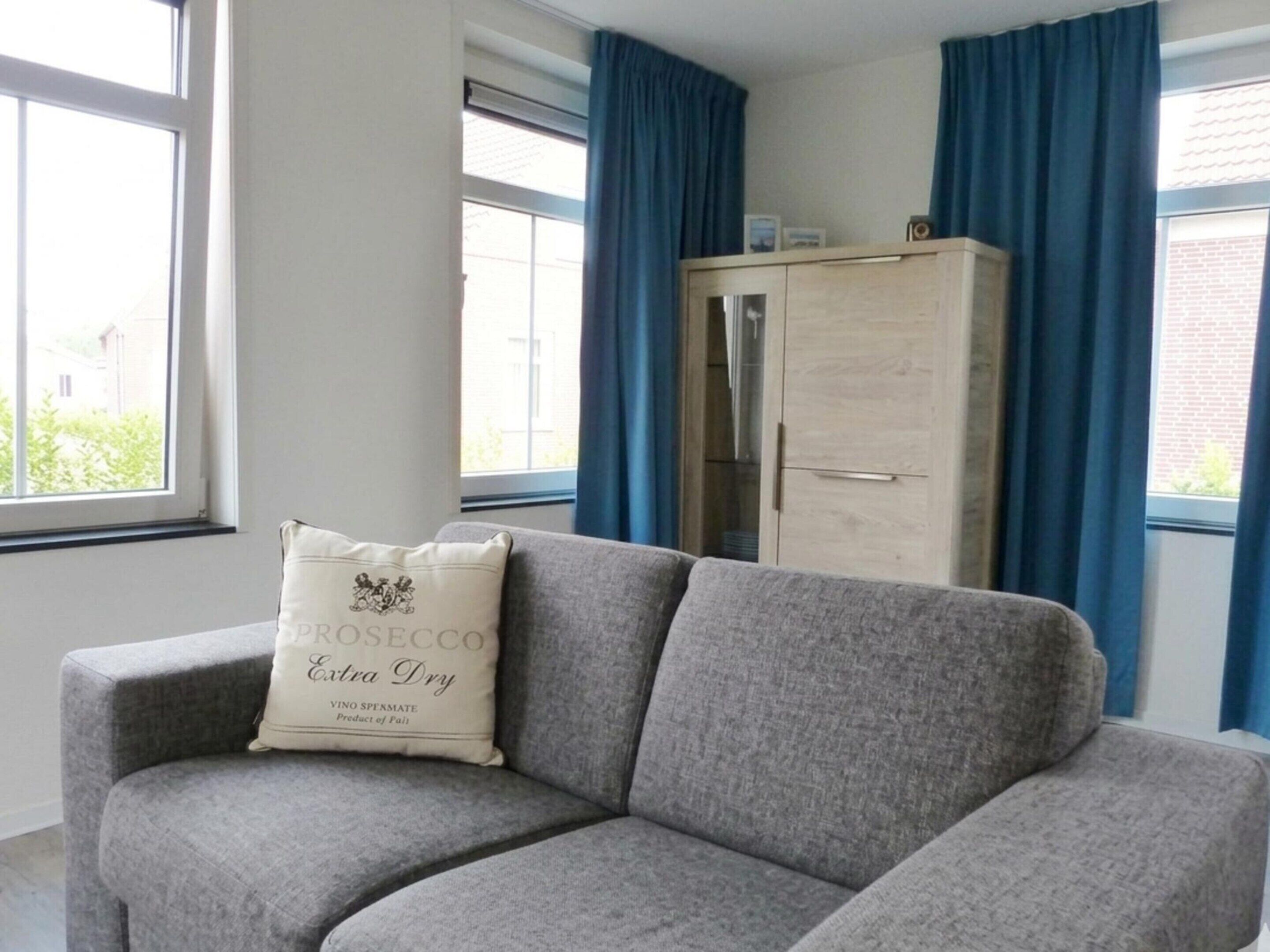 Un canapé gris avec un coussin portant l’inscription « ROSECCO Extra Dry », et un meuble en bois dans une pièce dotée de grandes fenêtres et de rideaux bleus.