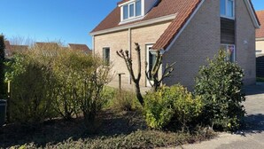 Une maison à deux étages avec un toit en pente, un petit garage attenant et un jardin agrémenté de divers arbustes et d’un cactus.