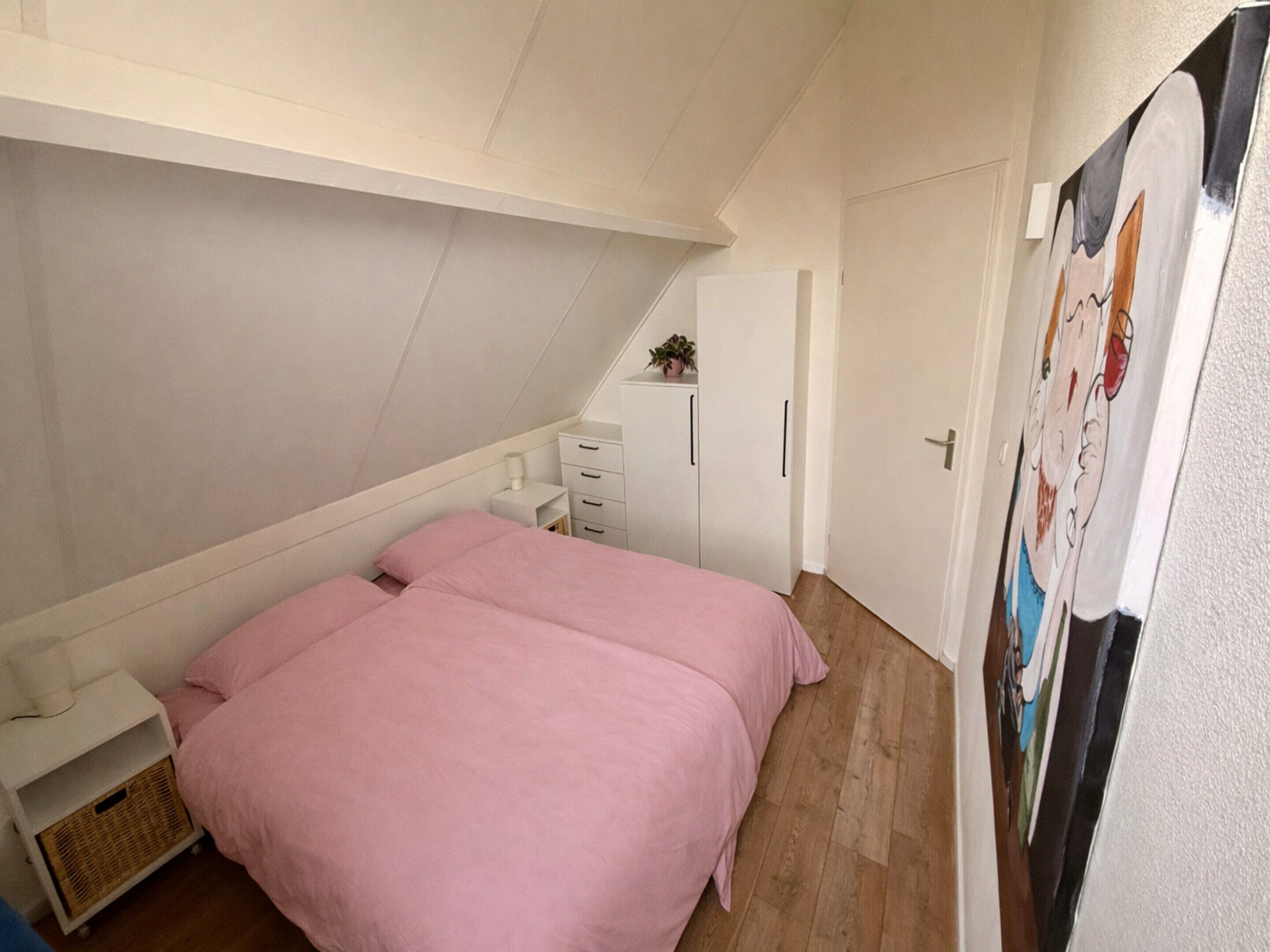 Une chambre avec un plafond en pente, un lit avec une literie rose, une armoire blanche et une porte ornée d’une affiche.