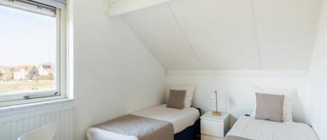 A small, single-room accommodation with two beds, a chair, a bedside table, and a window.